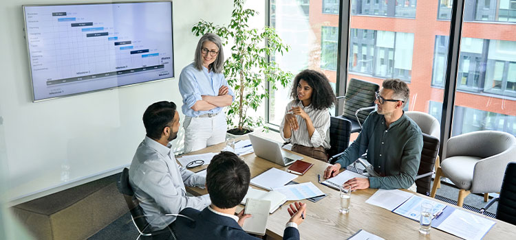 a woman standing in front of a wall screen with data talks to other professionals gathered around a table