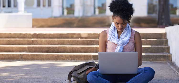 woman using laptop sitting on steps leading up to college campus building